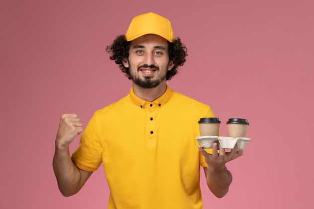 专业正面图身穿黄色制服和披风的男信使拿着棕色的咖啡杯在粉红色的墙上欢呼送货视野黄色