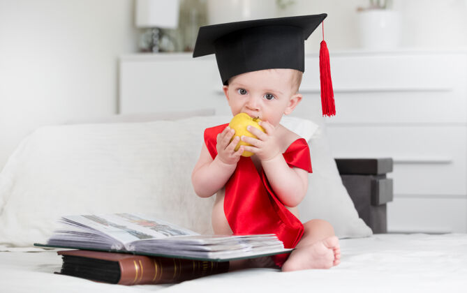 沙发戴着毕业帽的可爱男婴坐在书堆里咬着苹果的画像宝宝越来越聪明的概念卧室天才世代