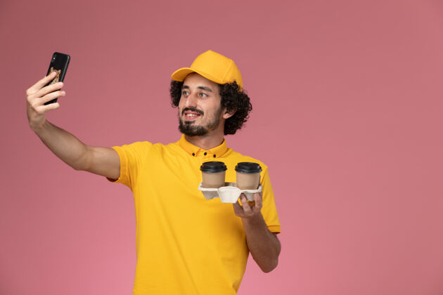 专业正面图身穿黄色制服 披风的男信使手持棕色咖啡杯在粉色墙上拍照男人黄色送货