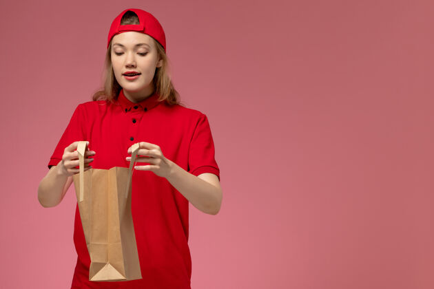 送货正面图：身穿红色制服 披风的年轻女快递员在粉色墙上拿着快递食品包正面手持包裹