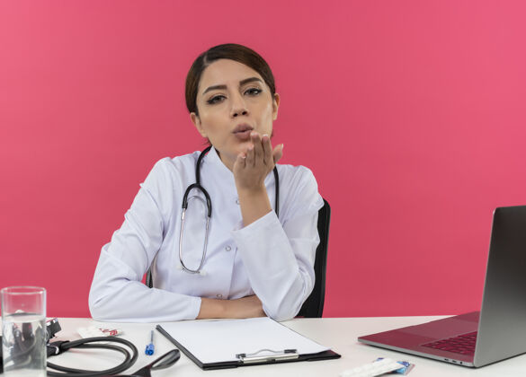 工具高兴的年轻女医生穿着医用长袍 听诊器 坐在办公桌上 用电脑和医疗工具 显示亲吻手势 并留有复印空间女性办公桌医疗