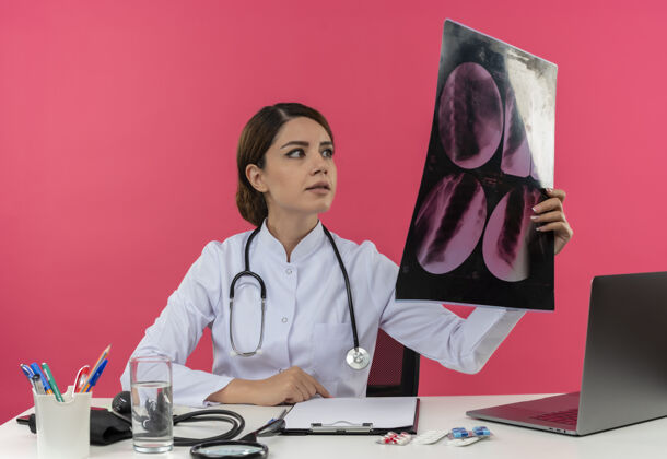 工具年轻的女医生穿着医用长袍 手持听诊器 坐在办公桌旁 拿着医疗工具 拿着电脑 看x光片 还有复印空间听诊器工作医疗